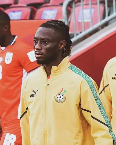 Gideon Mensah in a yellow Ghana jacket, his hair in locs, looking aside.